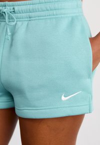 Artimas šviesiai mėlynų Nike sportinių šortų vaizdas su raišteliu prie juosmens ir ranka kišenėje tamsaus odos atspalvio žmogaus.