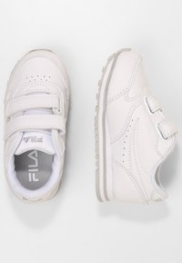 Scarpe sportive bianche con fasce in Velcro, parte superiore in pelle, colletto imbottito, perforazioni per traspirabilità e suola in gomma testurizzata.