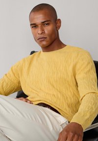 Gele kabelgebreide trui met een ronde halslijn en lange mouwen, gecombineerd met lichte broek; zittende positie, neutrale achtergrond.