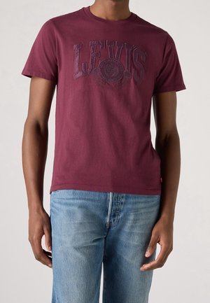 Burgundy cotton t-shirt featuring a raised, tonal "LEVI'S" logo on the chest. Short sleeves and round neckline. Paired with light blue jeans.