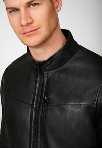 Black leather jacket with a stand collar, front zipper, and two zippered pockets. Smooth texture with a stitched design across the chest.