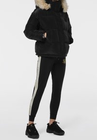 Person iført sort pufferjakke med pelskrave, sorte joggerbukser med beige sidestropper og sorte sneakers, står med hænderne i lommen.
