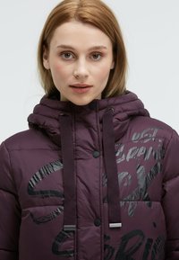 Purple puffer jacket with a high collar, featuring glossy black text patterns, adjustable hood, and button details. Smooth, quilted texture.