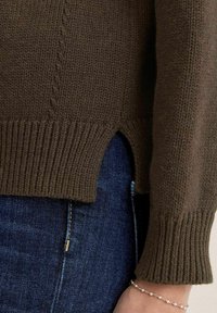 Brun strikket sweater med ribbet kant og manchet, sideslidser og et tekstureret mønster. Båret over mørke denimjeans med en synlig lynlås.