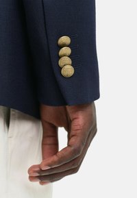 La manica della giacca da abito blu navy presenta quattro bottoni dorati in rilievo e un tessuto texturizzato. La mano è appoggiata di lato, mostrando i dettagli e il contrasto di colore.