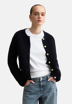 Donna che indossa un cardigan nero testurizzato con bottoni sopra una camicia bianca e jeans blu, in piedi con le mani nelle tasche contro uno sfondo semplice.