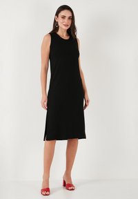 Black sleeveless dress with a straight silhouette, side slits, and a smooth texture. Worn with red heeled sandals.