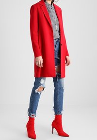 Rød lang frakke over en sort-hvid ternet skjorte, kombineret med slidte blå jeans og røde højhælede støvler. Glat tekstur, skræddersyet pasform.