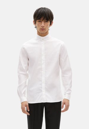 Camisa de rayas blancas con cuello alzado, que presenta un cierre de botones y mangas largas, hecha de un tejido suave. Combinada con pantalones negros.
