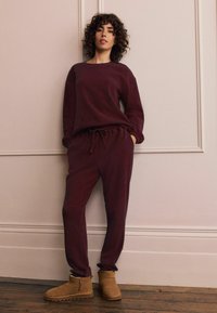 Bordó loungewear szett bordázott textúrával, amely egy lazán szabott, hosszú ujjú felsőt és zsinóros nadrágot tartalmaz, ébenbarna bokacsizmákkal párosítva.