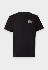 Melnā T-kreklā ar īsām piedurknēm, kuram uz kreisā krūtīm ir neliela balta "Stance Plumbing Service" logo un tālruņa numurs.