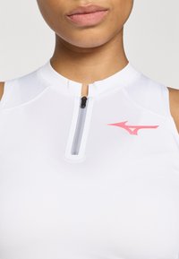 Top atletico bianco con colletto alto, dotato di una zip frontale e un accento del logo rosa sul lato. Tessuto liscio e leggero.