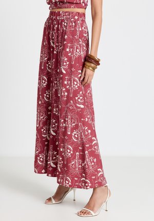 Frau trägt rote Hosen mit weitem Bein, bedruckt mit weißem Blumen- und Palmenmuster, beigefarbene Riemchen-High-Heels und Holzarmbänder am Handgelenk.