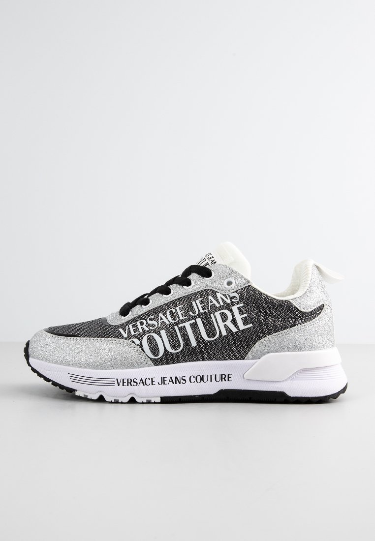 Zapatilla negra y plateada con suela blanca que presenta el texto "Versace Jeans Couture" en el lateral y la suela, cordones negros y detalles de tela texturizada.