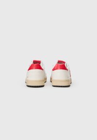 Hvite sneakers med røde hæl accenter, teksturert lær overdel, sort gummisåle og preget logo på hælen.