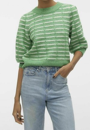 Person iført en grøn- og hvidstribet strikket sweater med puffede ærmer og lyseblå jeans med høj talje, hånd i lommen.