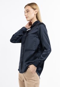 Camicia in satin blu navy con colletto classico, chiusura frontale con bottoni e taschino singolo sul petto; presenta maniche lunghe e una vestibilità rilassata.