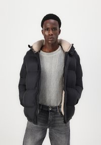 Black puffer jacket with a beige interior, featuring a high collar and zippered closure. Worn over a gray ribbed sweater and dark jeans.