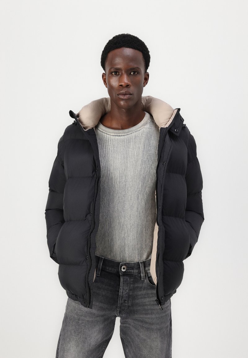 Schwarze Daunenjacke mit beigem Innenfutter, mit hohem Kragen und Reißverschluss. Getragen über einem grauen gerippten Pullover und dunklen Jeans.