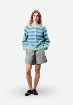 Femme portant un cardigan duveteux à rayures bleues et vert clair, un short ample gris, et des baskets noires, debout les mains dans les poches.