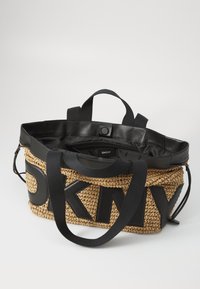 Grande bolsa tote com corpo de palha trançada e detalhes em couro preto, incluindo a alça. O interior revela um compartimento suave e forrado. Logotipo da DKNY exibido de forma proeminente.