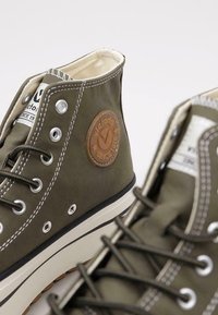 Zapatillas altas color verde oliva con suela de goma, que cuentan con costuras blancas, ojales de metal y un parche de logo marrón circular.