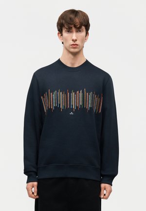 Νέος άνδρας που φοράει μαύρο φούτερ με πολύχρωμες κατακόρυφες γραμμές στο στήθος, στέκεται μπροστά σε ένα απλό φωτεινό φόντο.