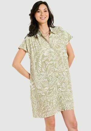 Naeratav naine kannab lühikeste varrukatega heledat rohelist nupuga kleiti valge lehemustriga ja kuldse ketiga kaelakeed, seisab käed selja taga.