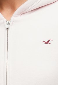 Ανοιχτό ροζ φούτερ με φερμουάρ, με απαλό ύφασμα, που διαθέτει μαρούν λογότυπο αετού στην αριστερή πλευρά του στήθους και ασημένιο φερμουάρ.