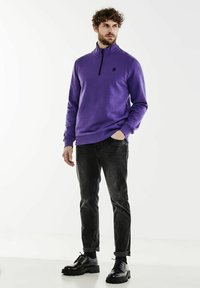 Purple zip-up sweatshirt with a high collar, paired with dark jeans and black shoes. Features a small logo on the chest. Smooth texture.
