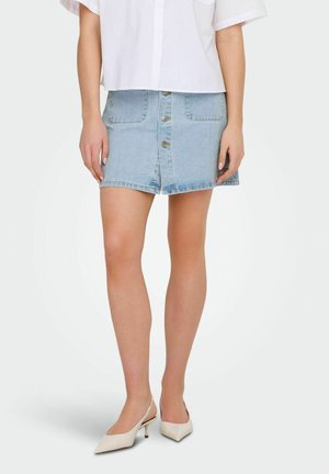 Femme portant une jupe en denim bleu clair avec boutons sur le devant, un chemisier blanc et des talons slingback blancs à bout pointu, debout sur un fond uni.