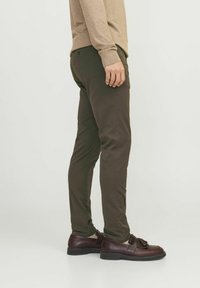 Schlank geschnittene, dunkelgrüne Hose aus glattem Stoff mit seitlichen Taschen und einem subtilen Glanz, kombiniert mit maronenfarbenen Slippern.