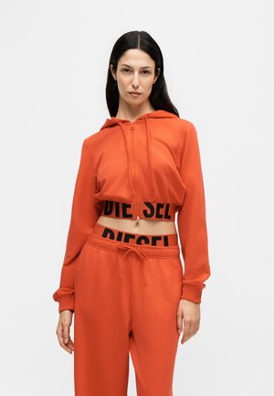 Kvinde iført orange cropped hættetrøje og matchende joggingbukser med sort Diesel-logo i taljen, står mod en ensfarvet lys baggrund.