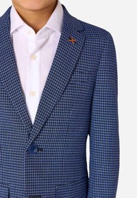 Blazer a motivo houndstooth blu e nero con un unico bottone, rever a punta e un'evidenza arancione sul rever. Camicia bianca sotto.