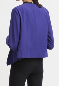 Lila långärmad topp med en slät textur, med hög halsringning och dragkedja i ryggen. Fållen är något kortare.