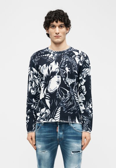Homme aux cheveux bouclés portant un pull à motif floral noir et blanc et un jean bleu déchiré, debout devant un fond uni.