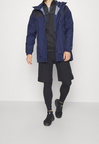 Granatowa wodoodporna kurtka z kapturem, czarne akcenty, elastyczne mankiety, w połączeniu z czarnymi szortami i czarnymi butami sportowymi.