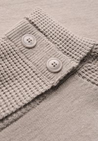 Ljust grått stickat tyg med en texturerad krage som har två runda knappar. Tyget är mjukt med en slät och ribbad struktur.