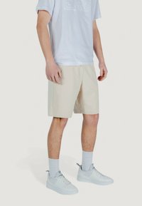 Shorts beige con tasche laterali, realizzati in tessuto leggero, indossati con una camicia bianca a maniche corte e sneakers bianche; vestibilità casual.