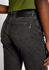 Black denim jeans with a slim fit, featuring stitched back pockets and a branded leather patch at the waist. Textured, slightly faded fabric.