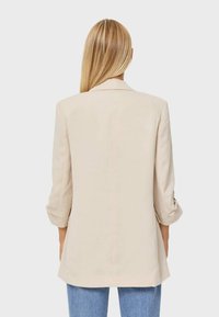 Beige blazer met opgerolde mouwen, een getailleerd ontwerp, een gladde textuur en een iets langere achterkant. Gecombineerd met lichtblauwe denim.