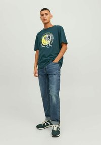 Camiseta gráfica de color verde oscuro con un logo circular que presenta un esqueleto y texto, combinada con jeans azul claro y zapatillas verde oscuro.