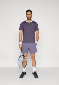 Άνδρας που στέκεται κρατώντας ρακέτα τένις, φορώντας μωβ αθλητική μπλούζα και σορτς, λευκά περιβραχιόνια, λευκές κάλτσες και μαύρα αθλητικά παπούτσια σε απλό φόντο.