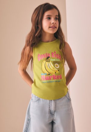 Ung pige med langt brunt hår iført en grøn ærmeløs top med bananer og farverig tekst, kombineret med lyse denimbukser, stående indendørs.
