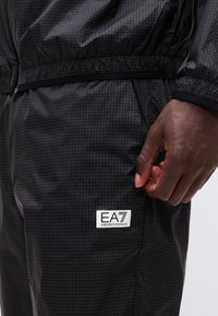 Κοντινό πλάνο ενός σκούρου σακακιού και παντελονιού με σχέδιο πλέγματος, που φέρει ένα λευκό έμβλημα με το λογότυπο EA7 Emporio Armani κοντά στην τσέπη και ένα χέρι ακουμπισμένο στο πλάι.