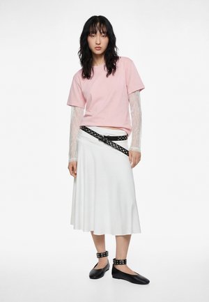 Donna che indossa una maglietta rosa a maniche corte sopra maniche lunghe in pizzo bianco, gonna midi bianca, cintura nera con borchie e ballerine nere con cinturino alla caviglia.