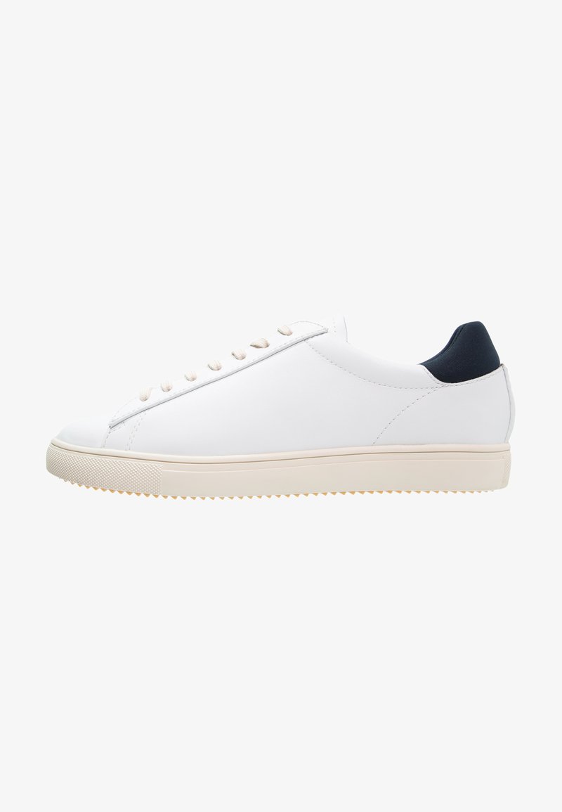 White sneaker with a smooth texture, featuring beige laces and a navy heel accent. The sole is light beige with a textured surface.