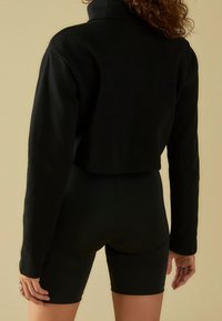 Sweat-shirt noir court avec col montant, manches longues et tissu texturé, associé à un short de cycliste noir, offrant une coupe ajustée.