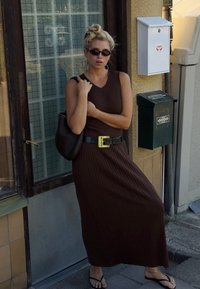 Robe maxi côtelée marron avec un décolleté en V, cintrée à la taille par une ceinture noire avec une boucle jaune. Portée avec des tongs noires et des lunettes de soleil.