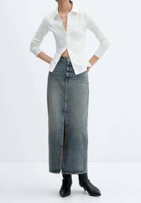 Bílá košile s krátkým rukávem, zkrácená do pasu, zkombinovaná s dlouhou, rovnou šedou denimovou sukni s předním rozparkem a černými kotníkovými botami.